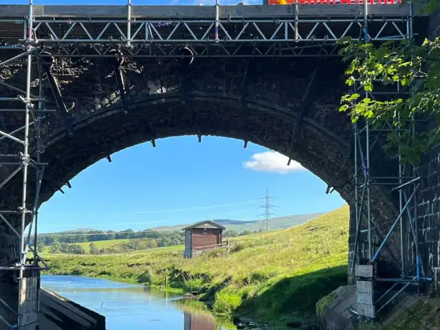 Scaffold with a beamed working platform under a stone arch bridge over a river